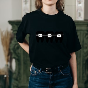 May include: A black t-shirt with a graphic of three teeth with braces. The braces are white and the teeth are black.