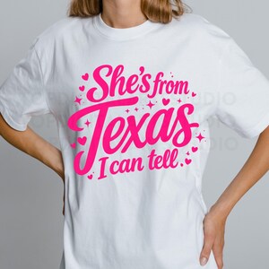 K&ouml;nnte beinhalten: Wei&szlig;es T-Shirt mit dem Text "She's from Texas I can tell" in leuchtendem Pink. Das Design beinhaltet Herzen und Sterne. Das T-Shirt hat einen l&auml;ssigen Stil.