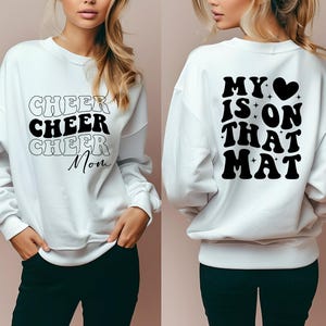 May include: White sweatshirt with black text. The front says "Cheer Cheer Cheer Mom" and the back says "My heart is on that mat".