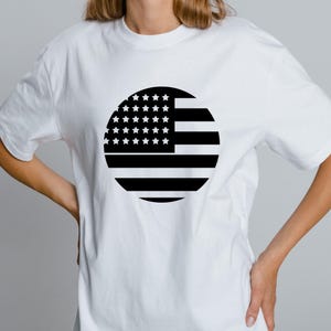May include: White t-shirt featuring a black and white circular graphic of the American flag. The design includes stars and stripes, representing the United States of America. The shirt is a casual, everyday wear item.