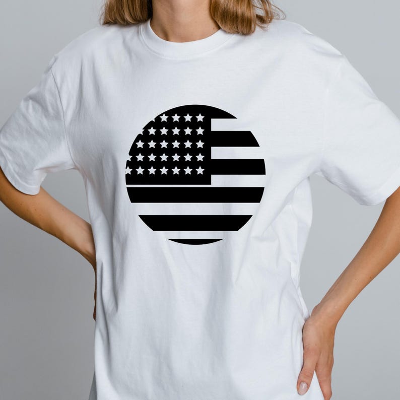 May include: White t-shirt featuring a black and white circular graphic of the American flag. The design includes stars and stripes, representing the United States of America. The shirt is a casual, everyday wear item.