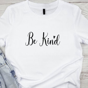 Pode incluir: Camiseta branca com o texto "Be Kind" em letras cursivas pretas com um pequeno s&iacute;mbolo de cora&ccedil;&atilde;o.