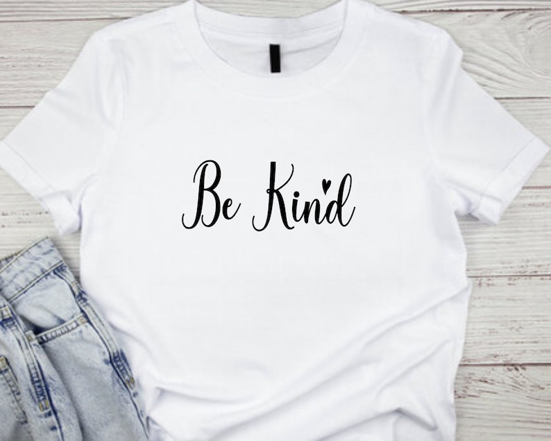 Pode incluir: Camiseta branca com o texto "Be Kind" em letras cursivas pretas com um pequeno s&iacute;mbolo de cora&ccedil;&atilde;o.