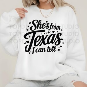 K&ouml;nnte beinhalten: Wei&szlig;es Sweatshirt mit dem Text "She's from Texas I can tell" in schwarzer Schrift. Das Design umfasst kleine Herzen und Sterne. Das Sweatshirt ist ein l&auml;ssiger Stil.