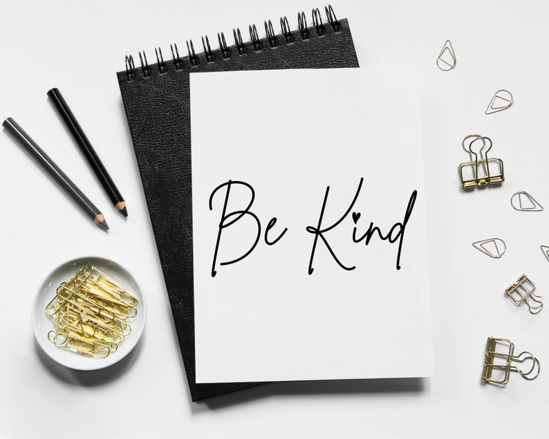 Pode incluir: Uma folha de papel branca com as palavras "Be Kind" escritas em tinta preta em um caderno preto com clipes de pasta dourados e l&aacute;pis sobre uma superf&iacute;cie branca.