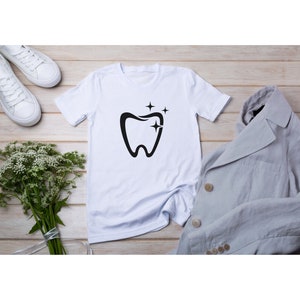 May include: White t-shirt with a black tooth graphic and three stars above it. The shirt is laid flat on a wooden surface with a gray blazer and white sneakers.