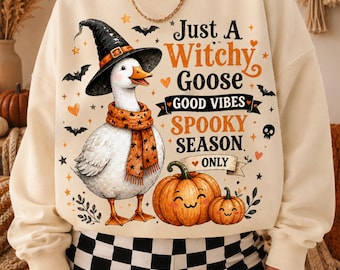 Puede incluir: Una sudadera de cuello redondo color crema con el gráfico de una oca blanca que lleva un sombrero de bruja negro y una bufanda naranja con estrellas. El texto dice Just A Witchy Goose GOOD VIBES SPOOKY SEASON ONLY, junto a calabazas y murciélagos.