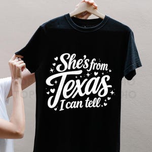 K&ouml;nnte beinhalten: Schwarzes T-Shirt mit dem wei&szlig;en Text "She's from Texas I can tell." Der Text ist in einer Schreibschrift mit dekorativen Herzen und Sternen gehalten. Das T-Shirt h&auml;ngt an einem Holzb&uuml;gel.