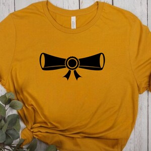 以下が含まれることがあります： マスタードイエローのTシャツに、リボンで結ばれた卒業証書の黒いシルエットが描かれています。