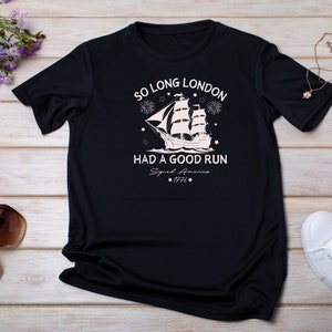May include: Black t-shirt with a white graphic of a ship sailing away with fireworks and stars. The text reads "So Long London Had A Good Run Signed America 1776".