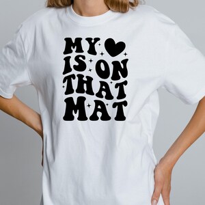 May include: White t-shirt with black text that reads "MY &hearts; IS ON THAT MAT" in a retro font.