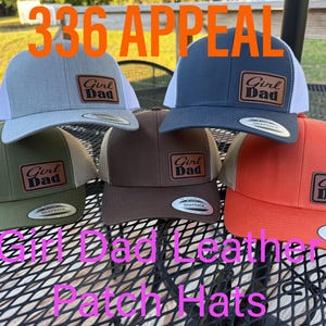 May include: Five snapback hats in gray, dark blue, olive green, brown, and orange, each featuring a brown leather patch with the text Girl Dad. The hats have mesh backs and a structured crown design with a flat brim and a circular sticker on the bill.
