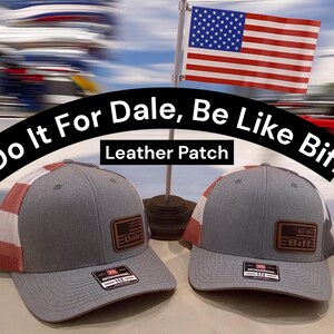 May include: Two grey trucker hats with mesh backs featuring red and white stripes. Each hat has a brown leather patch on the front. One patch reads Do It For Dale and the other reads Be Like Biff. Both hats have a black Richardson 112 sticker on the brim.
