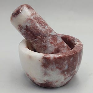 May include: A mortar and pestle set made of marble. The mortar is a bowl shape, and the pestle is cylindrical. The marble has a mix of white and red hues, creating a natural, patterned appearance. This kitchen tool is ideal for grinding herbs and spices.