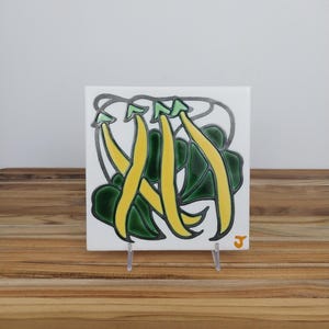 May include: A square ceramic tile featuring a raised relief design of three yellow bean pods with light green tips and dark green leaves, outlined in dark gray. The tile has a white background and a small orange letter J in the bottom right corner.