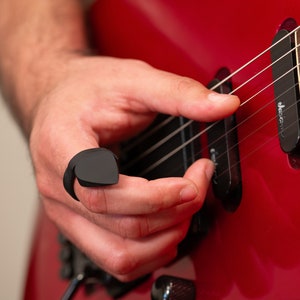 May include: A person's hand playing a red electric guitar with a black pick on their finger. The guitar has a black pick-up and the text "Jackson" on the body.