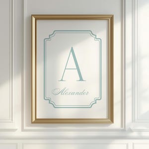Könnte beinhalten: Ein gerahmter Druck mit dem hellblauen Buchstaben "A" und dem Namen "Alexander" in Schreibschrift. Das Kunstwerk befindet sich vor einem weißen Hintergrund mit einem hellblauen dekorativen Rahmen, goldfarben gerahmt. Der Druck hängt an einer weißen Wand.