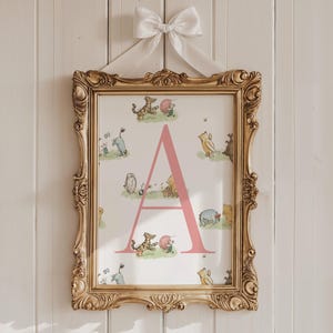 May include: A framed print with the letter "A" in pink, and illustrations of Winnie the Pooh characters. The print is in an ornate gold frame, suspended by a white ribbon bow. The background is a white wooden wall, typical of a nursery or child's room.