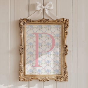 May include: A framed print with the letter "P" in pink, set against a floral pattern of blue and pink flowers. The print is in a gold ornate frame and is suspended by a white ribbon and bow. The artwork hangs on a white wooden wall.