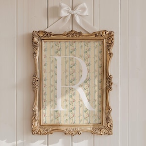 May include: A framed print with a gold ornate frame, featuring a floral and striped background with the letter "R" in white. The frame is suspended by a white ribbon tied in a bow at the top. The background is a light yellow with blue stripes and floral accents.