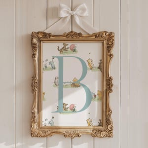 Puede incluir: Un estampado enmarcado con la letra "B" en azul claro y un tema de Winnie the Pooh. El marco dorado adornado está suspendido por un lazo de cinta blanca. El fondo es una pared de madera blanca.