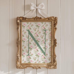 May include: A framed print with a gold ornate frame, featuring a floral pattern with pink roses and a large green letter "N". The print is hung with a white ribbon and bow against a white wooden background.