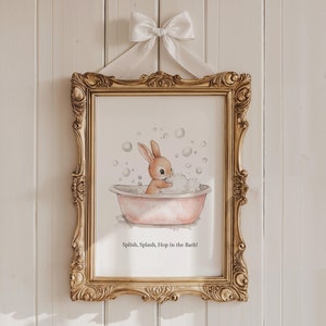 May include: A framed print featuring a watercolor illustration of a bunny taking a bath in a pink tub filled with bubbles. The gold frame is ornate and is suspended by a white ribbon and bow. The text at the bottom reads: "Splish, Splash, Hop in the Bath!"