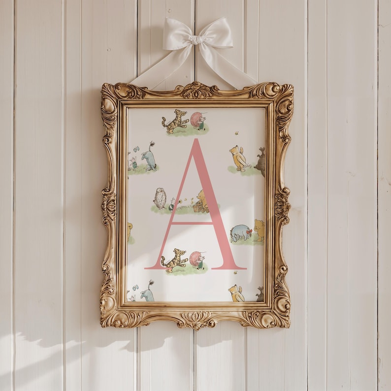 May include: A framed print featuring the letter "A" in pink, surrounded by illustrations of Winnie the Pooh characters. The print is set in an ornate gold frame and is suspended by a white ribbon tied in a bow. The background is a white wooden wall.