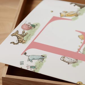 May include: A white print featuring the letter "F" in pink, with illustrations of cartoon characters. The print includes a tiger, a donkey, a pig, and other characters in a whimsical design. The print is set inside a wooden box.