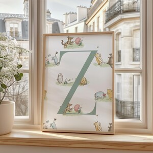 May include: A framed print featuring the letter "Z" in a soft green hue, adorned with illustrations of Winnie the Pooh characters. The artwork is set against a white background and framed in light wood, displayed on a windowsill.