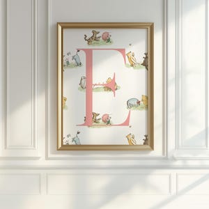 May include: A framed print featuring a pink letter "E" with illustrations of Winnie the Pooh characters. The artwork is set against a white background and framed in a gold-colored frame. The print is mounted on a white wall.