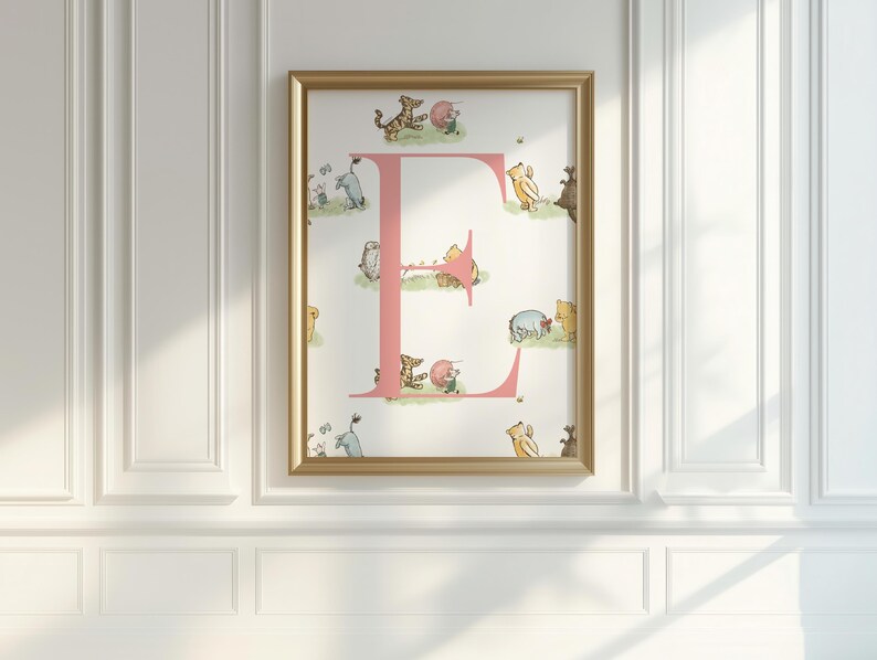 May include: A framed print featuring a pink letter "E" with illustrations of Winnie the Pooh characters. The artwork is set against a white background and framed in a gold-colored frame. The print is mounted on a white wall.