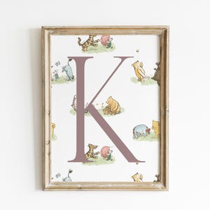 May include: A framed print featuring the letter "K" in a mauve color, surrounded by illustrations of Winnie the Pooh characters. The artwork has a light beige wooden frame and is set against a white background.