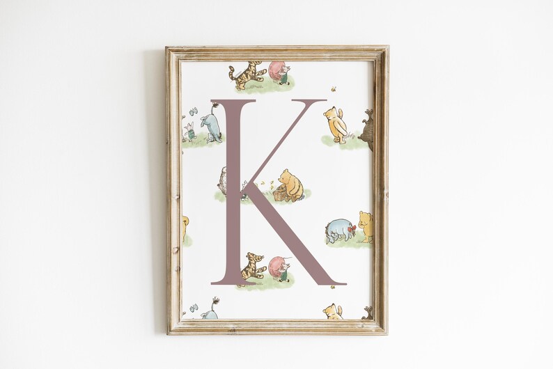 May include: A framed print featuring the letter "K" in a mauve color, surrounded by illustrations of Winnie the Pooh characters. The artwork has a light beige wooden frame and is set against a white background.