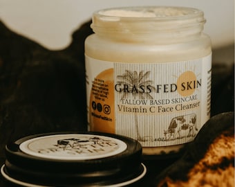 Può includere: Un barattolo bianco con un'etichetta nera che recita "Grass Fed Skin Tallow Based Skincare Vitamin C Face Cleanser". Il barattolo è appoggiato su una superficie marrone. L'etichetta presenta anche una palma e un sole.