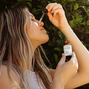 May include: A woman applies a serum to her face. The bottle is labeled "Primal Radiance Serum" and is made by Grass Fed Skin.