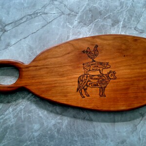 May include: A wooden cutting board with a unique handle shaped like a figure eight. The board is engraved with a diagram of a cow, pig, and chicken, showing the different cuts of meat.