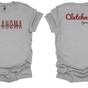 K&ouml;nnte beinhalten: Hellgraues T-Shirt mit den W&ouml;rtern "OKLAHOMA SOONERS" auf der Vorderseite in Burgunderrot und Schwarz. Auf der R&uuml;ckseite des Shirts steht das Wort "Clutchness" in burgunderroter Schrift, darunter "-Gasso". Das Shirt hat kurze &Auml;rmel.