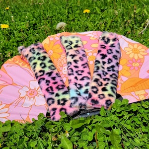 May include: Three fuzzy leopard print fabric wrist cuffs in pink, blue, and green. The cuffs are laid out on a pink and white floral fabric background.