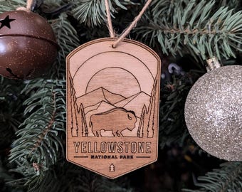 Puede incluir: Adorno de madera con forma de insignia de guardabosques con una silueta de bisonte y montañas. El adorno es marrón y tiene el texto "Yellowstone National Park".