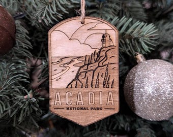 Puede incluir: Un adorno de madera con un diseño grabado con láser de un faro y una costa. El adorno es marrón y tiene el texto "Acadia National Park".