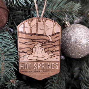 May include: Wooden Christmas ornament with a laser-cut design of a waterfall and trees. The ornament reads "HOT SPRINGS NATIONAL PARK" in a rectangular shape. The ornament hangs from a natural twine string.