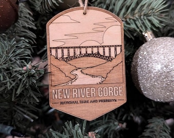 Puede incluir: Un adorno de madera con una imagen grabada con láser de un puente sobre un río. El adorno tiene forma de etiqueta y tiene el texto "NEW RIVER GORGE NATIONAL PARK AND PRESERVE".