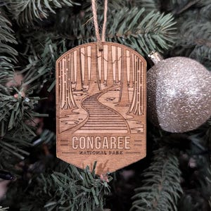 May include: A wooden Christmas ornament featuring a laser-engraved design of a path through a forest. The ornament is brown and has the text "Congaree National Park" on it.