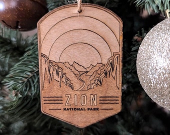 Puede incluir: Un adorno de madera con una imagen grabada con láser de un paisaje de cañón. El adorno es de color marrón y tiene el texto "Zion National Park".