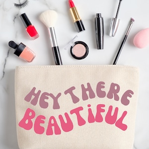 May include: A beige canvas makeup bag with the text "Hey There Beautiful" in pink and purple lettering. The bag is open and filled with makeup products, including a red lipstick, a black mascara, a black eyeliner, a pink blush, a clear nail polish bottle, and a makeup brush.
