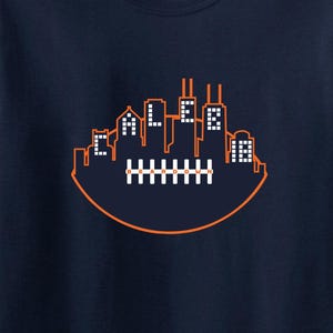 May include: Navy blue t-shirt with an orange football-shaped design containing a cityscape. The cityscape includes the text "CALEB" in white block letters. The shirt is made of a soft, comfortable material.