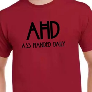 Peut inclure: T-shirt bordeaux avec le texte noir "AHD" au-dessus de "ASS HANDED DAILY". Le t-shirt à col rond et à manches courtes présente le texte centré sur le devant.