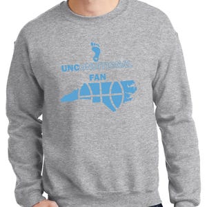 K&ouml;nnte beinhalten: Ein graues Sweatshirt mit einem hellblauen Aufdruck. Der Aufdruck zeigt einen Fu&szlig;abdruck, die Worte "UNCONDITIONAL FAN" und eine stilisierte Abbildung des Bundesstaates North Carolina mit einem Basketball-Design.