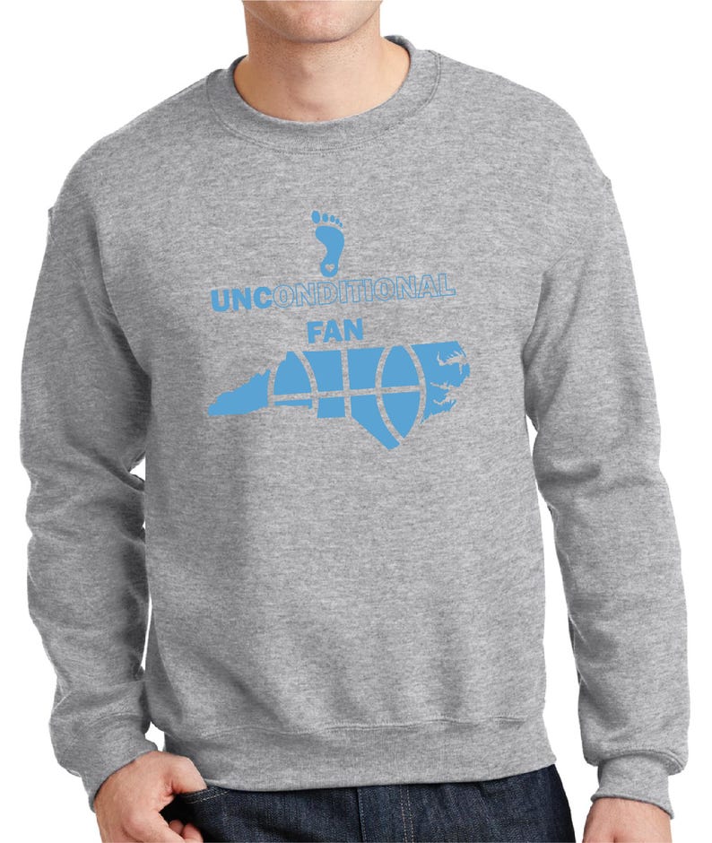 K&ouml;nnte beinhalten: Ein graues Sweatshirt mit einem hellblauen Aufdruck. Der Aufdruck zeigt einen Fu&szlig;abdruck, die Worte "UNCONDITIONAL FAN" und eine stilisierte Abbildung des Bundesstaates North Carolina mit einem Basketball-Design.